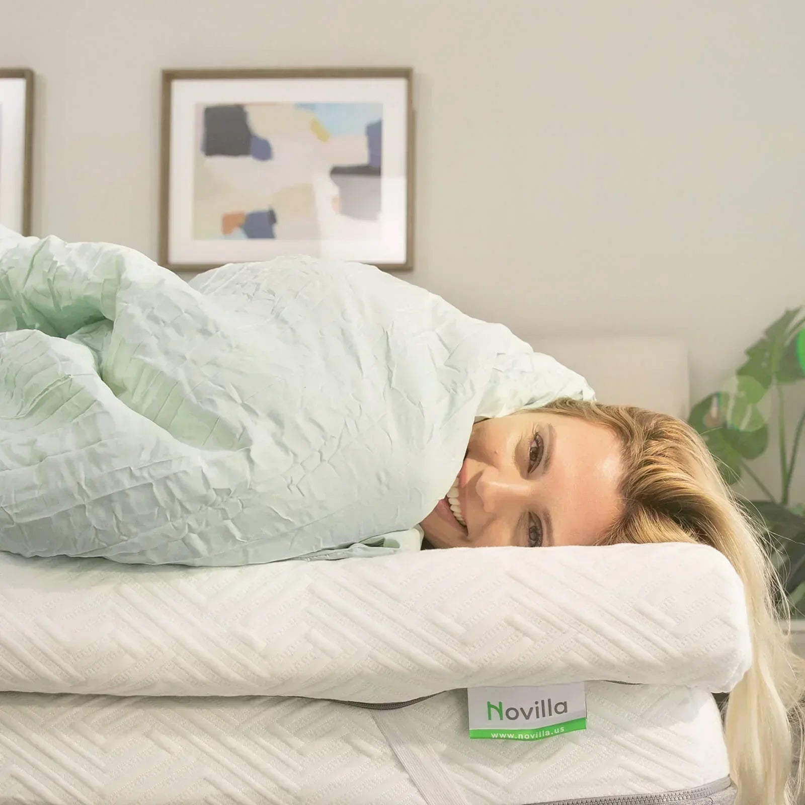 A smiling woman wrapped in a mint green comforter, lying on a Novilla mattress topper in a cozy bedroom