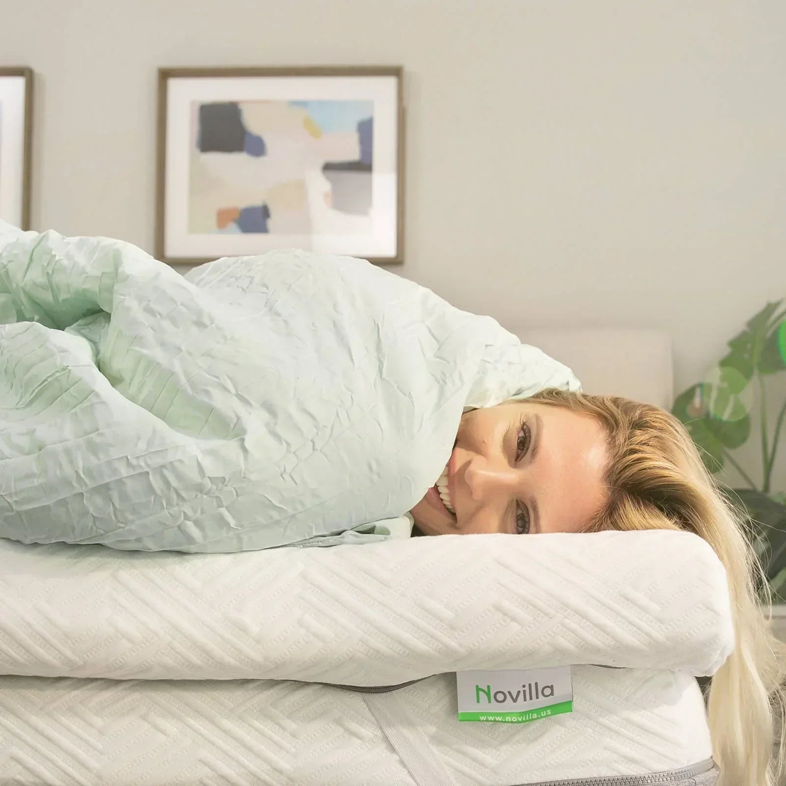 A smiling woman wrapped in a mint green comforter, lying on a Novilla mattress topper in a cozy bedroom